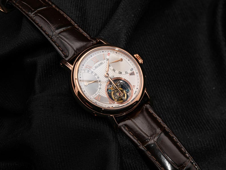 May include: A luxury wristwatch with a rose gold case and a white dial. The watch features a brown leather strap and a visible tourbillon movement. The dial includes sub-dials and Roman numeral hour markers. The watch is displayed on a black surface.