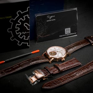 May include: A gold-toned wristwatch with a white face and brown leather band, accompanied by an extra band and a tool. The watch has visible internal mechanisms. A black instruction booklet and a card with the text "Sugess Seestern" are also present.
