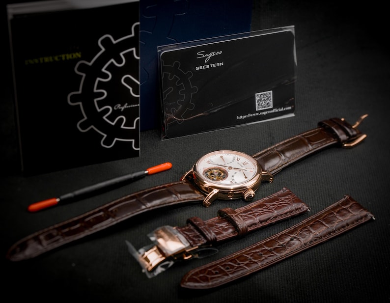 May include: A gold-toned wristwatch with a white face and brown leather band, accompanied by an extra band and a tool. The watch has visible internal mechanisms. A black instruction booklet and a card with the text "Sugess Seestern" are also present.