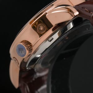 May include: Close-up of a rose gold wristwatch with a brown leather strap. The watch features a detailed face with a blue accent on the crown. The watch has a polished finish and a classic design.