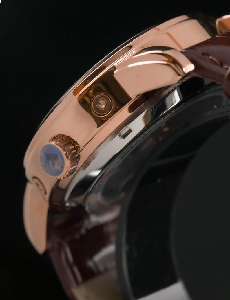 May include: Close-up of a rose gold wristwatch with a brown leather strap. The watch features a detailed face with a blue accent on the crown. The watch has a polished finish and a classic design.