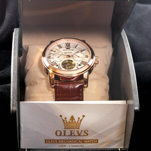 May include: A gold-toned OLEVS mechanical watch with a brown leather band, presented in a black box. The watch face features Roman numerals and a visible inner mechanism. The box includes a user's manual.