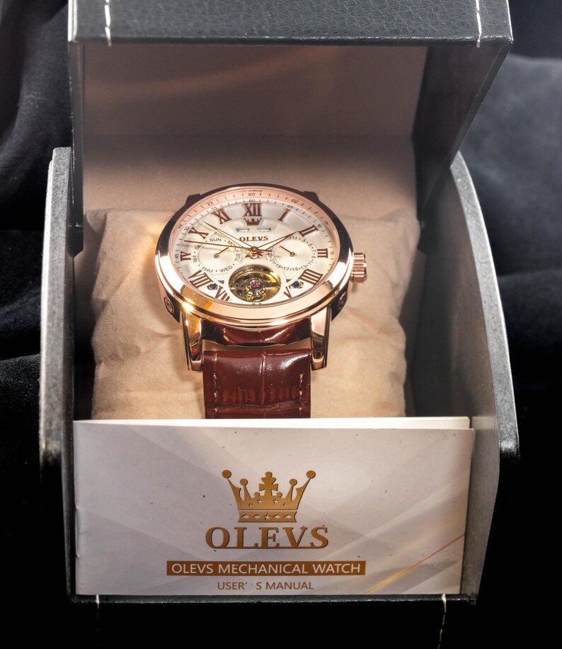 May include: A gold-toned OLEVS mechanical watch with a brown leather band, presented in a black box. The watch face features Roman numerals and a visible inner mechanism. The box includes a user's manual.
