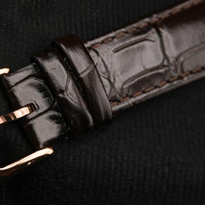 May include: Close-up of a brown leather watch strap with a rose gold buckle. The strap has a textured, crocodile-skin pattern. The buckle is rectangular with a pin closure. The watch strap is on a black background.