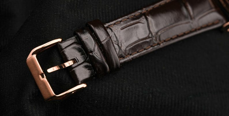 May include: Close-up of a brown leather watch strap with a rose gold buckle. The strap has a textured, crocodile-skin pattern. The buckle is rectangular with a pin closure. The watch strap is on a black background.