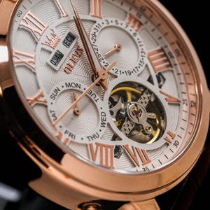 May include: Close-up of a rose gold wristwatch with a white face and Roman numeral hour markers. The watch features a calendar display, day of the week indicators, and a visible mechanical movement. The watch has a leather strap.