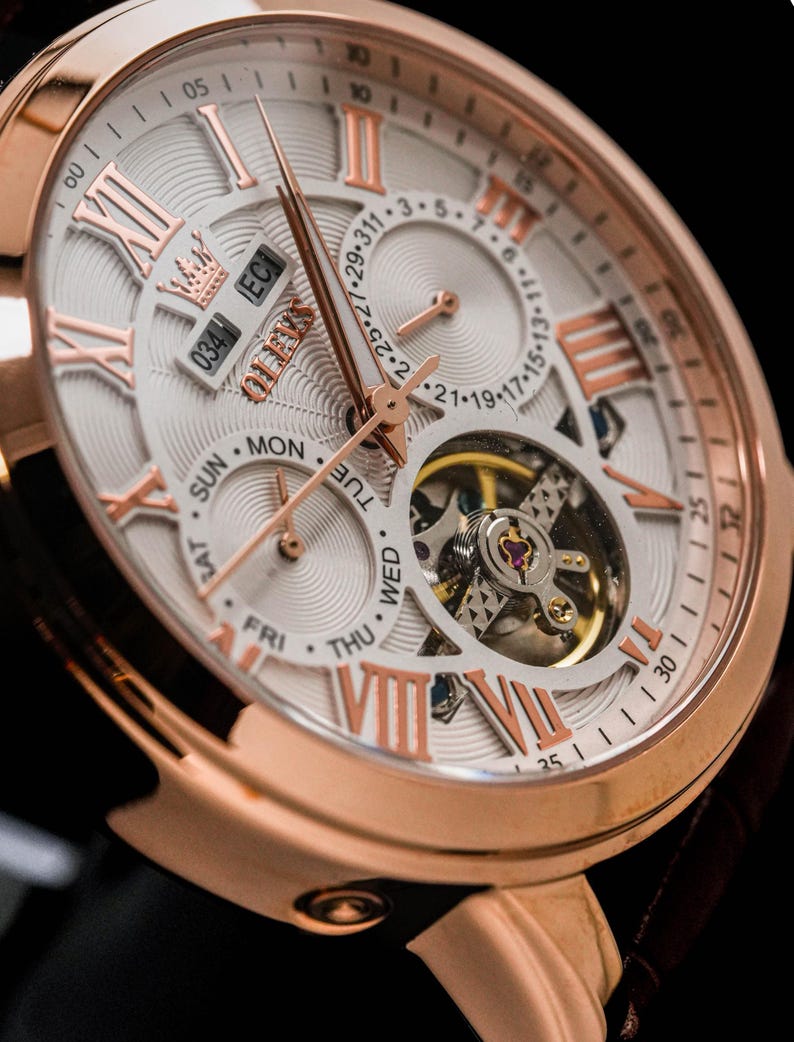 May include: Close-up of a rose gold wristwatch with a white face and Roman numeral hour markers. The watch features a calendar display, day of the week indicators, and a visible mechanical movement. The watch has a leather strap.