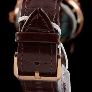 May include: Close-up of a brown leather watch strap with a gold-colored buckle. The strap has a textured, crocodile-like pattern. A white tag is attached to the strap. The watch face is visible in the background.