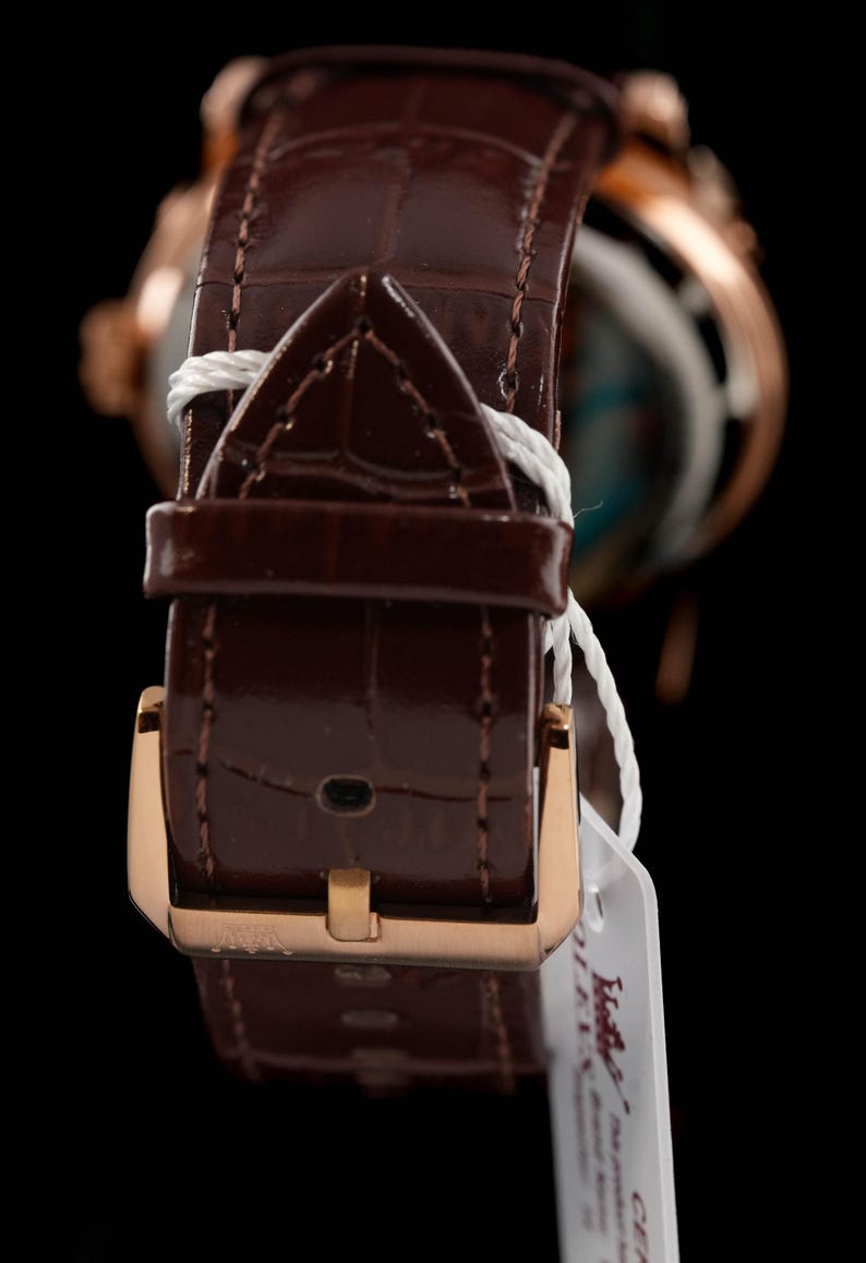 May include: Close-up of a brown leather watch strap with a gold-colored buckle. The strap has a textured, crocodile-like pattern. A white tag is attached to the strap. The watch face is visible in the background.