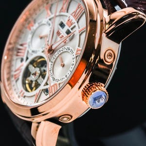 May include: Close-up of a rose gold wristwatch with a white face and Roman numeral hour markers. The watch features a brown leather band and a blue crown. The watch face displays multiple sub-dials and intricate mechanical details.