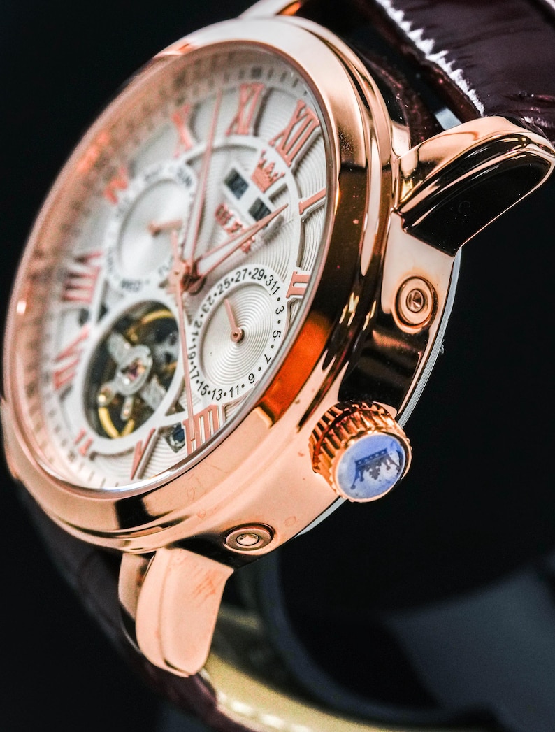 May include: Close-up of a rose gold wristwatch with a white face and Roman numeral hour markers. The watch features a brown leather band and a blue crown. The watch face displays multiple sub-dials and intricate mechanical details.
