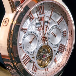 May include: Close-up of a rose gold wristwatch with a silver face. The watch features Roman numeral hour markers, day and date subdials, and a visible escapement. The watch has a black leather strap.