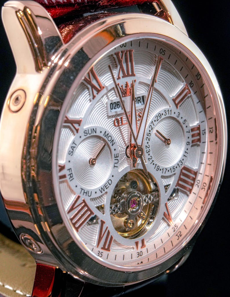 May include: Close-up of a rose gold wristwatch with a silver face. The watch features Roman numeral hour markers, day and date subdials, and a visible escapement. The watch has a black leather strap.