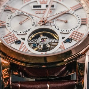May include: Close-up of a rose gold wristwatch with a white face and Roman numeral hour markers. The watch features a visible escapement and a brown leather strap. The face displays the days of the week and the date.