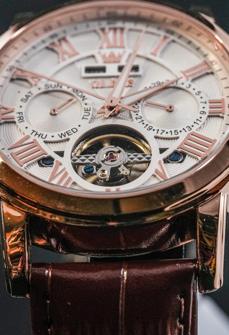 May include: Close-up of a rose gold wristwatch with a white face and Roman numeral hour markers. The watch features a visible escapement and a brown leather strap. The face displays the days of the week and the date.