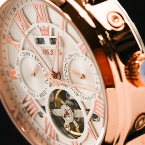 May include: Close-up of a rose gold wristwatch with a white face. The watch features Roman numeral hour markers, day and date displays, and intricate inner workings visible through a circular window. The watch hands and numerals are in a contrasting rose gold.