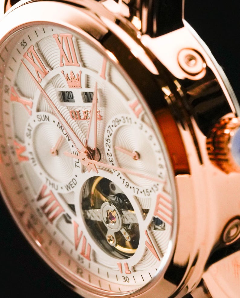 May include: Close-up of a rose gold wristwatch with a white face. The watch features Roman numeral hour markers, day and date displays, and intricate inner workings visible through a circular window. The watch hands and numerals are in a contrasting rose gold.