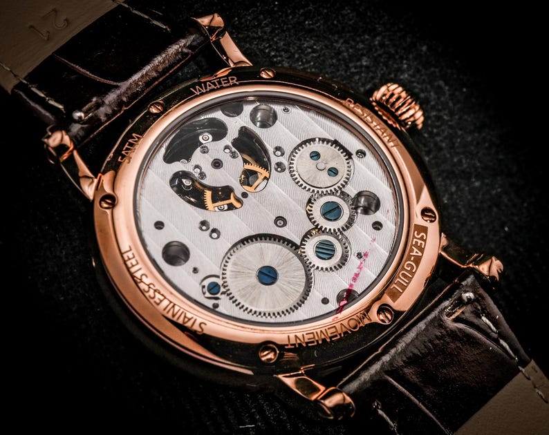 May include: Close-up of a rose gold wristwatch with a transparent back, showcasing the intricate mechanical movement. The watch has a brown leather strap and the words "WATER", "STAINLESS STEEL", and "SEA-GULL" are visible.
