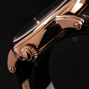 May include: Close-up of a rose gold wristwatch with a dark brown leather strap. The watch features a round face with a clear crystal and a textured crown. The watch's metal casing has a polished finish, reflecting light.