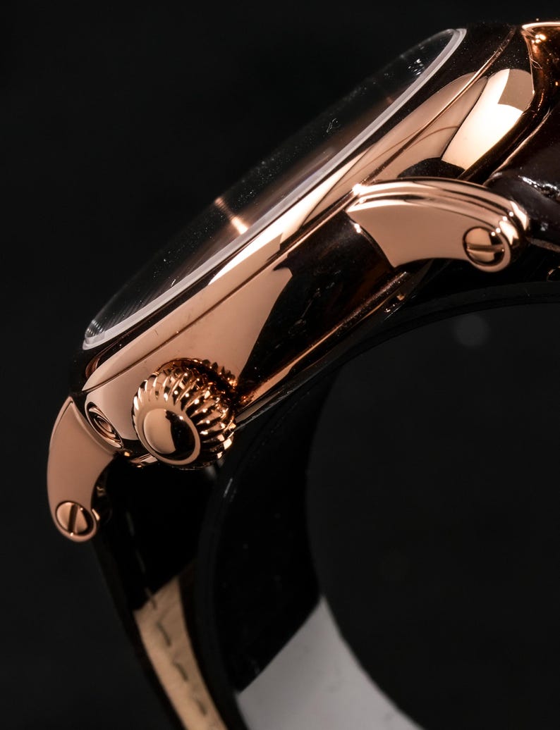 May include: Close-up of a rose gold wristwatch with a dark brown leather strap. The watch features a round face with a clear crystal and a textured crown. The watch's metal casing has a polished finish, reflecting light.