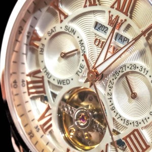 May include: Close-up of a gold and white wristwatch. The watch face features Roman numerals, day and date indicators, and a visible mechanical movement. The hands and accents are gold-toned, set against a white background.
