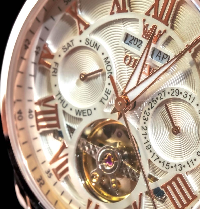 May include: Close-up of a gold and white wristwatch. The watch face features Roman numerals, day and date indicators, and a visible mechanical movement. The hands and accents are gold-toned, set against a white background.