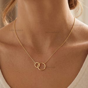 May include: A gold-toned necklace featuring a delicate chain and two interlocking circular rings as a central pendant. The necklace rests against the neck, showcasing a simple design with a polished metallic finish and a secure clasp mechanism.