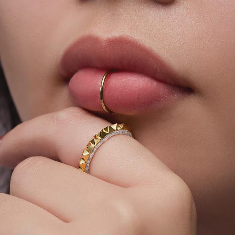May include: Close-up of a person's lips and hand adorned with jewelry. A gold ring is in the lip, and a gold and silver ring with pyramid-shaped accents is on the finger. The skin tone is fair, and the lips are a natural pink.