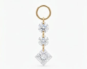 Puede incluir: Un charm dorado con tres piedras preciosas transparentes. Dos diamantes de talla redonda están conectados por una fina cadena a un diamante de talla cuadrada en la base. El charm tiene un lazo circular en la parte superior.