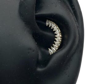 May include: A curved ear cuff featuring a row of clear, teardrop-shaped gemstones. The gemstones are set in a gold-colored metal, creating a sparkling, elegant accessory. The cuff is shown on a black ear model.