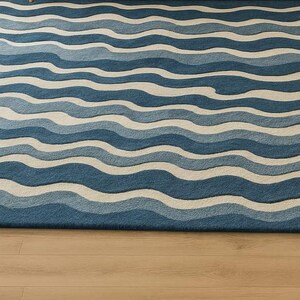 Pu&ograve; includere: Un tappeto rettangolare con un motivo a onde in tonalit&agrave; di blu e bianco sporco. Il design del tappeto ricorda le onde dell'oceano, creando un'estetica rilassante e visivamente accattivante. Il tappeto &egrave; posizionato su un pavimento in legno chiaro.