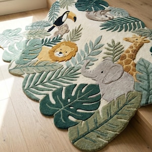 May include: An irregularly shaped area rug featuring a jungle theme with a cream background. The rug displays tufted illustrations of a toucan, a monkey, a lion, a sloth, an elephant, and a giraffe surrounded by various green tropical leaves.