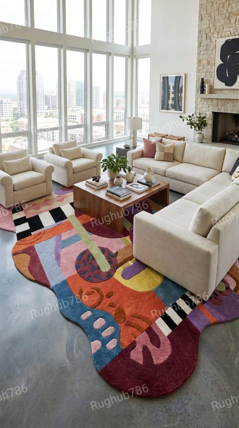 May include: A modern living room featuring a colorful, abstract area rug with a wavy edge. The rug's design incorporates various shapes and colors, including orange, blue, and purple. Beige sofas and armchairs surround a wooden coffee table, creating a comfortable seating area.