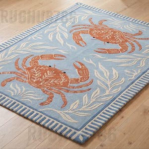 Op de afbeelding: Een rechthoekig vloerkleed met een lichtblauwe achtergrond, twee oranje krabben en witte zeewierpatronen. Het kleed heeft een gestreepte rand in lichtblauw en wit. De tekst RUGHUB is zichtbaar in de linker boven- en benedenhoek.