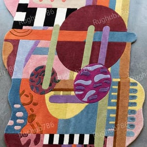 May include: Abstract area rug with a colorful, geometric design. The rug features a mix of shapes and colors, including pink, black, white, orange, blue, purple, and green. The rug has a unique, irregular shape.