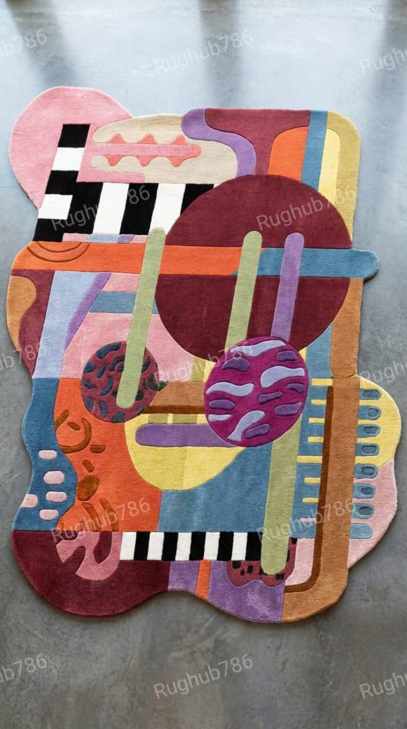 May include: Abstract area rug with a colorful, geometric design. The rug features a mix of shapes and colors, including pink, black, white, orange, blue, purple, and green. The rug has a unique, irregular shape.