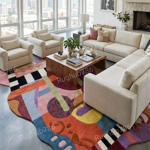 May include: A modern living room featuring a colorful, abstract area rug with a unique shape. The rug incorporates shades of red, orange, blue, and purple. The room includes cream-colored sofas, armchairs, and a wooden coffee table, all set against a backdrop of large windows.