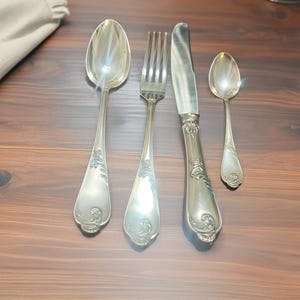 Può includere: Set di posate d'argento composto da un cucchiaio, una forchetta, un coltello e un cucchiaino. Le posate hanno manici decorativi e sono disposte su una superficie di legno scuro. Un panno piegato è visibile nell'angolo in alto a sinistra.