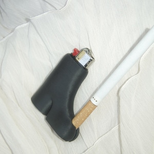 Puede incluir: Un estuche negro para encendedor con un botón rojo y un encendedor de metal plateado está junto a un cigarrillo blanco con filtro beige. Los artículos están sobre una superficie blanca texturizada.