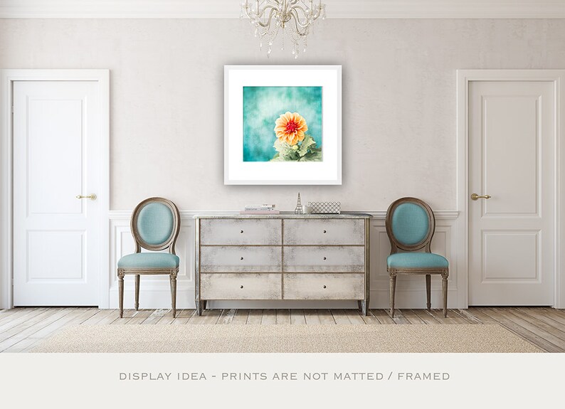 May include: A framed floral print featuring a vibrant orange and yellow dahlia against a teal and turquoise background. The artwork is displayed above a distressed gray chest of drawers, flanked by two teal upholstered chairs. The text at the bottom reads: DISPLAY IDEA - PRINTS ARE NOT MATTED / FRAMED.