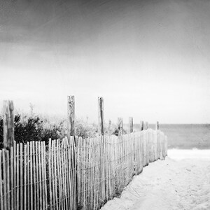 Puede incluir: Fotograf&iacute;a en blanco y negro de una escena de playa. Una valla de madera desgastada recorre la orilla arenosa, con el oc&eacute;ano visible a lo lejos bajo un cielo nublado. La valla est&aacute; hecha de listones de madera verticales.