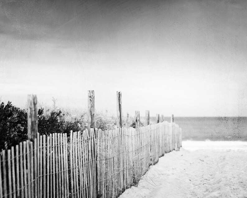 Puede incluir: Fotograf&iacute;a en blanco y negro de una escena de playa. Una valla de madera desgastada recorre la orilla arenosa, con el oc&eacute;ano visible a lo lejos bajo un cielo nublado. La valla est&aacute; hecha de listones de madera verticales.