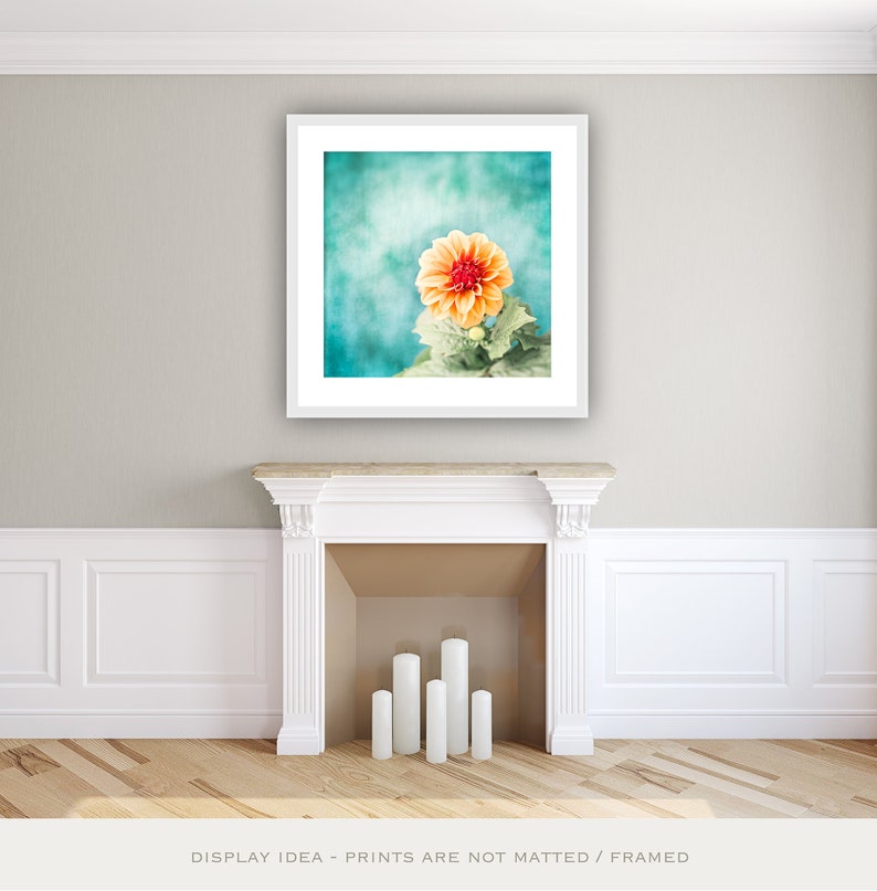 May include: A framed print featuring a close-up of an orange dahlia flower with a red center, set against a teal background. The flower is surrounded by green leaves. The print is displayed above a white fireplace with candles.