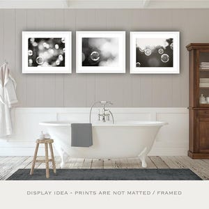 May include: Three framed monochrome bubble prints above a white clawfoot bathtub. A wooden cabinet and a small wooden stool are also in the bathroom. A grey bath mat and towel complete the scene.