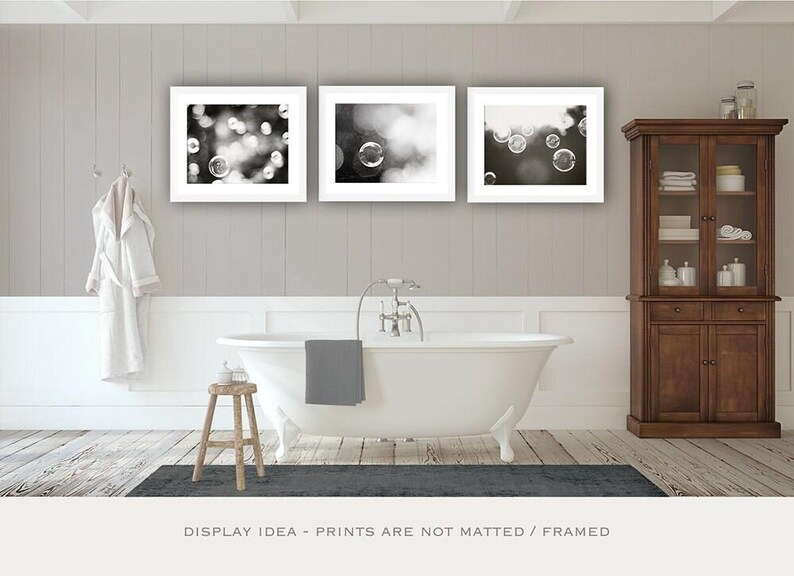 May include: Three framed monochrome bubble prints above a white clawfoot bathtub. A wooden cabinet and a small wooden stool are also in the bathroom. A grey bath mat and towel complete the scene.