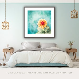 May include: A framed print featuring a close-up of an orange dahlia flower against a teal background. The image is displayed above a bed with a white comforter, flanked by wooden nightstands and hanging gold lanterns. The text at the bottom reads: DISPLAY IDEA - PRINTS ARE NOT MATTED / FRAMED.