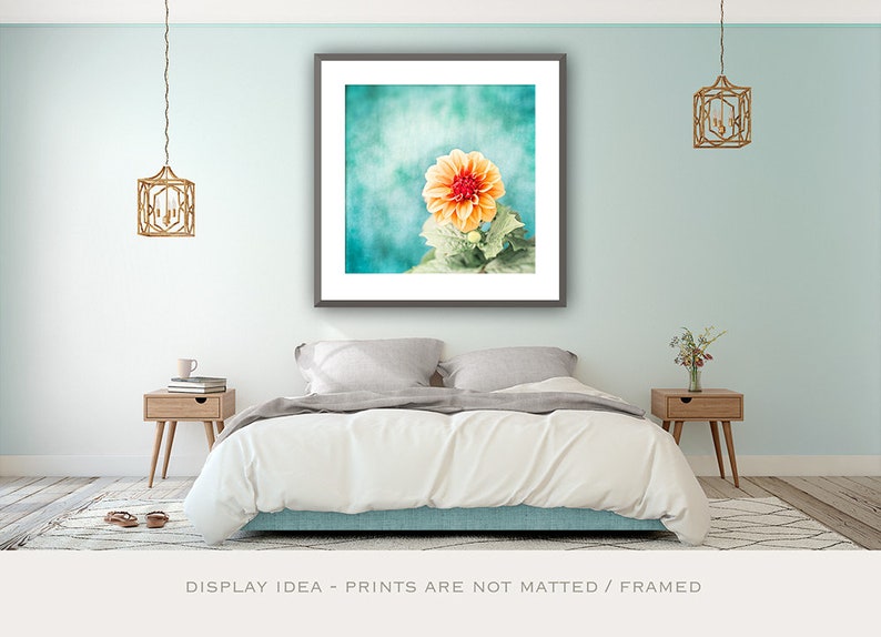 May include: A framed print featuring a close-up of an orange dahlia flower against a teal background. The image is displayed above a bed with a white comforter, flanked by wooden nightstands and hanging gold lanterns. The text at the bottom reads: DISPLAY IDEA - PRINTS ARE NOT MATTED / FRAMED.