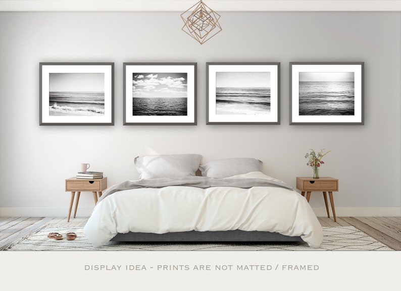 Puede incluir: Cuatro impresiones enmarcadas en blanco y negro de paisajes marinos, exhibidas sobre una cama. Las impresiones representan varias escenas costeras, incluyendo olas y mar abierto. El dormitorio presenta una paleta de colores neutros con mesitas de noche de madera.