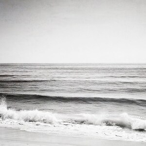 Puede incluir: Fotograf&iacute;a en blanco y negro del oc&eacute;ano. La imagen captura el mar con suaves olas que rompen en la orilla. El cielo es de un gris p&aacute;lido y el agua de un tono m&aacute;s oscuro, creando una escena costera serena y minimalista.