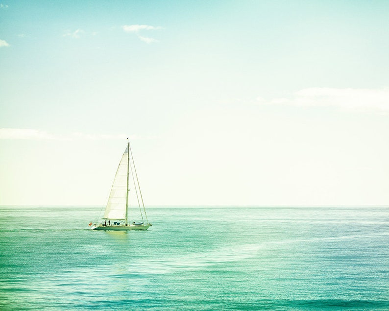 May include: A white sailboat with a tall mast and billowing sails glides across the turquoise ocean under a clear, light blue sky. The image evokes a sense of tranquility and adventure, with the boat as the central focus.